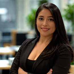 Woman with long dark hair, wearing a black blazer, arms crossed, smiling in an office setting.