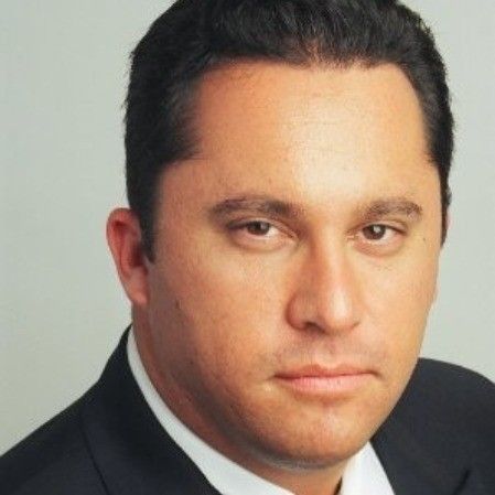 Man in dark suit, looking directly at the camera with a neutral expression.