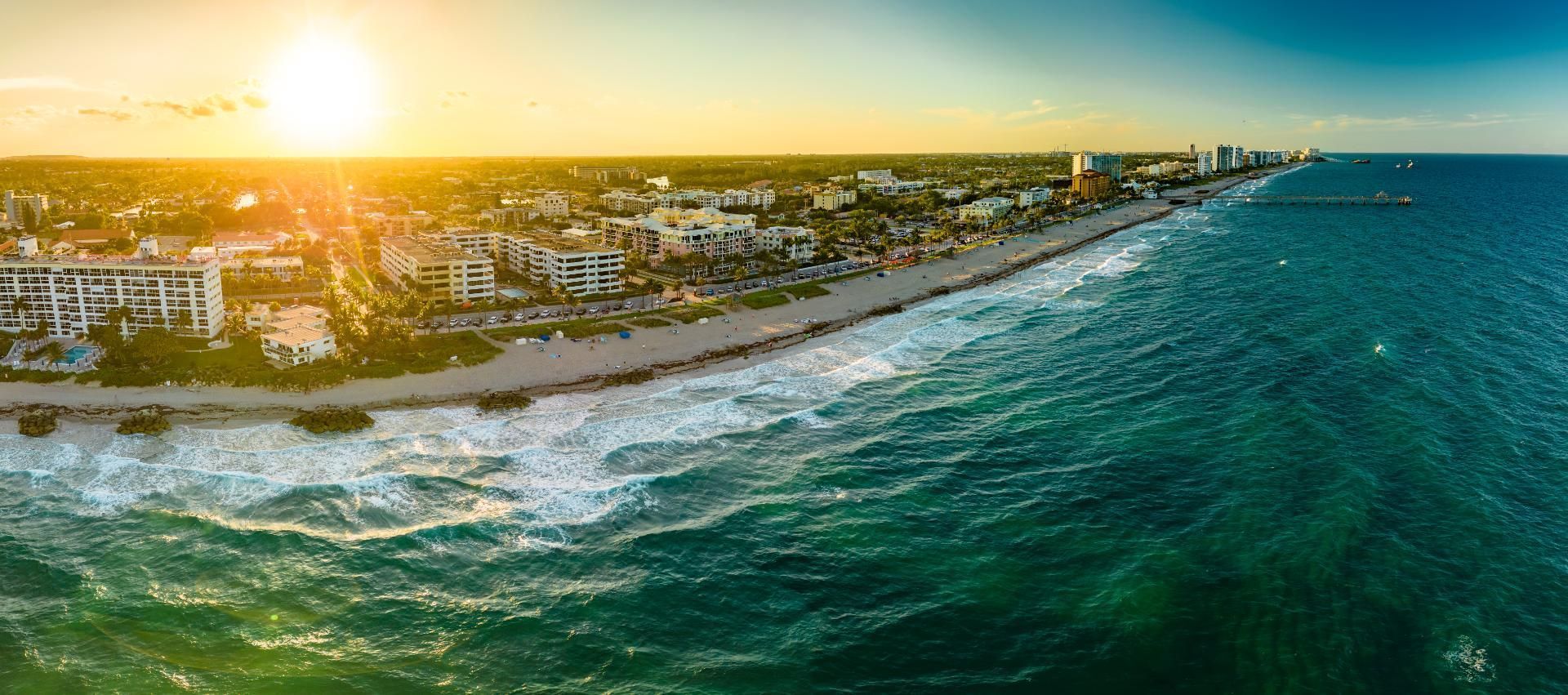 Aerial view of a coastal city at sunset, with golden light over the buildings, beach, and ocean.