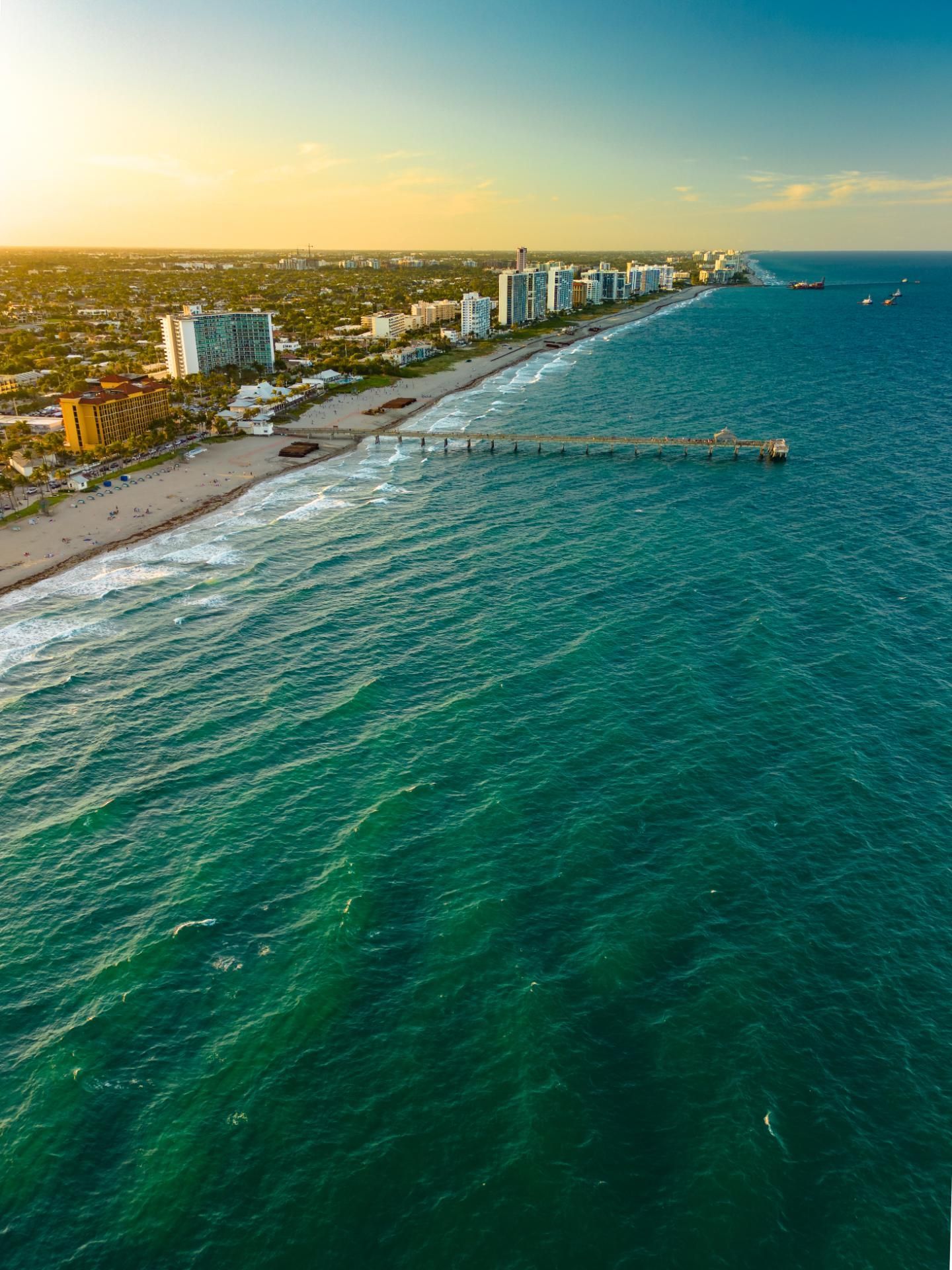 Aerial view of a beach with turquoise water, pier, buildings, and a golden sunset sky.