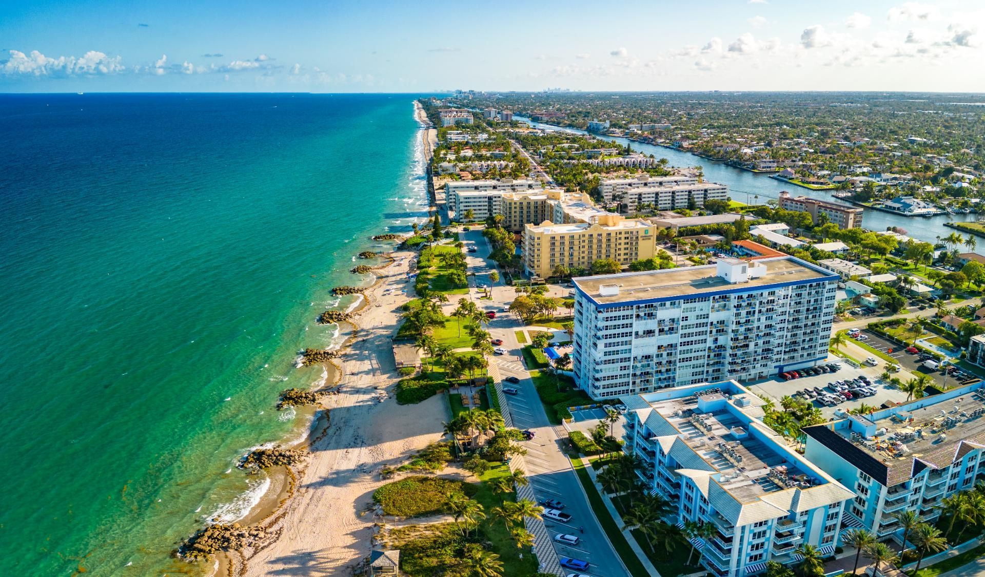 Aerial view of a sandy beach next to turquoise ocean, and urban buildings lining the coast.