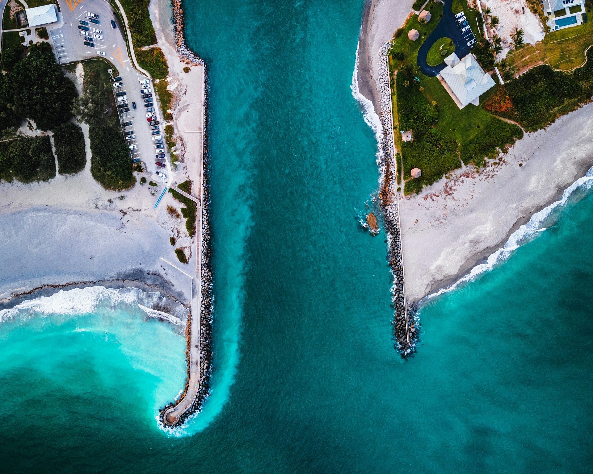 Aerial view of a turquoise ocean inlet between rocky jetties, with sandy beaches and buildings.