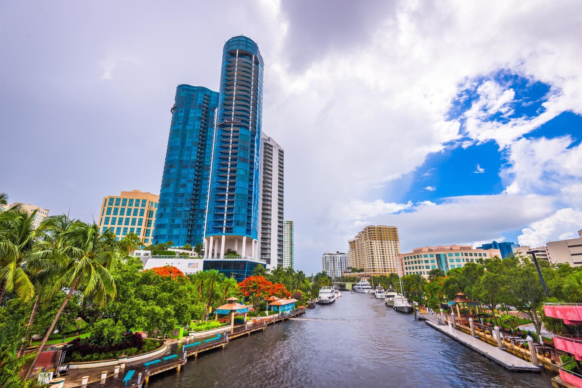 Cityscape with blue skyscraper, river, boats, and lush greenery under a cloudy sky.