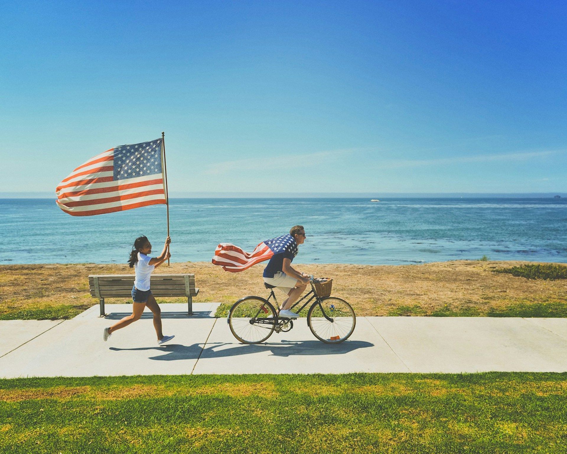 Girl running with American flag, man riding bike with flag draped as cape, seaside path.