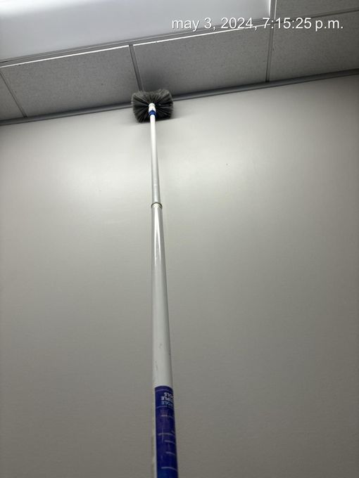 Pole brush reaching up to clean a ceiling corner with a black brush head on a gray wall.