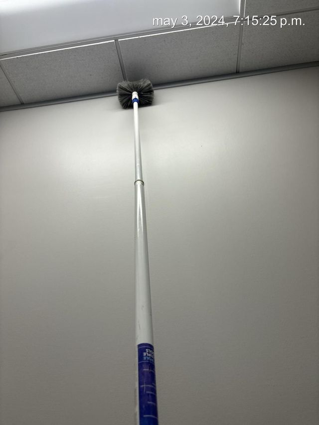 Pole brush reaching up to clean a ceiling corner with a black brush head on a gray wall.