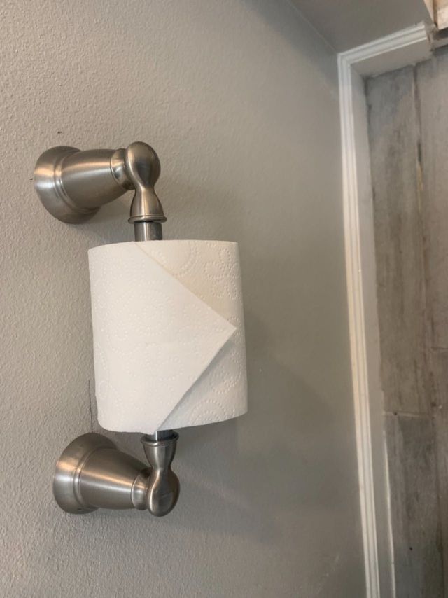 Wall-mounted metal toilet paper holder with a roll of white paper in a bathroom.