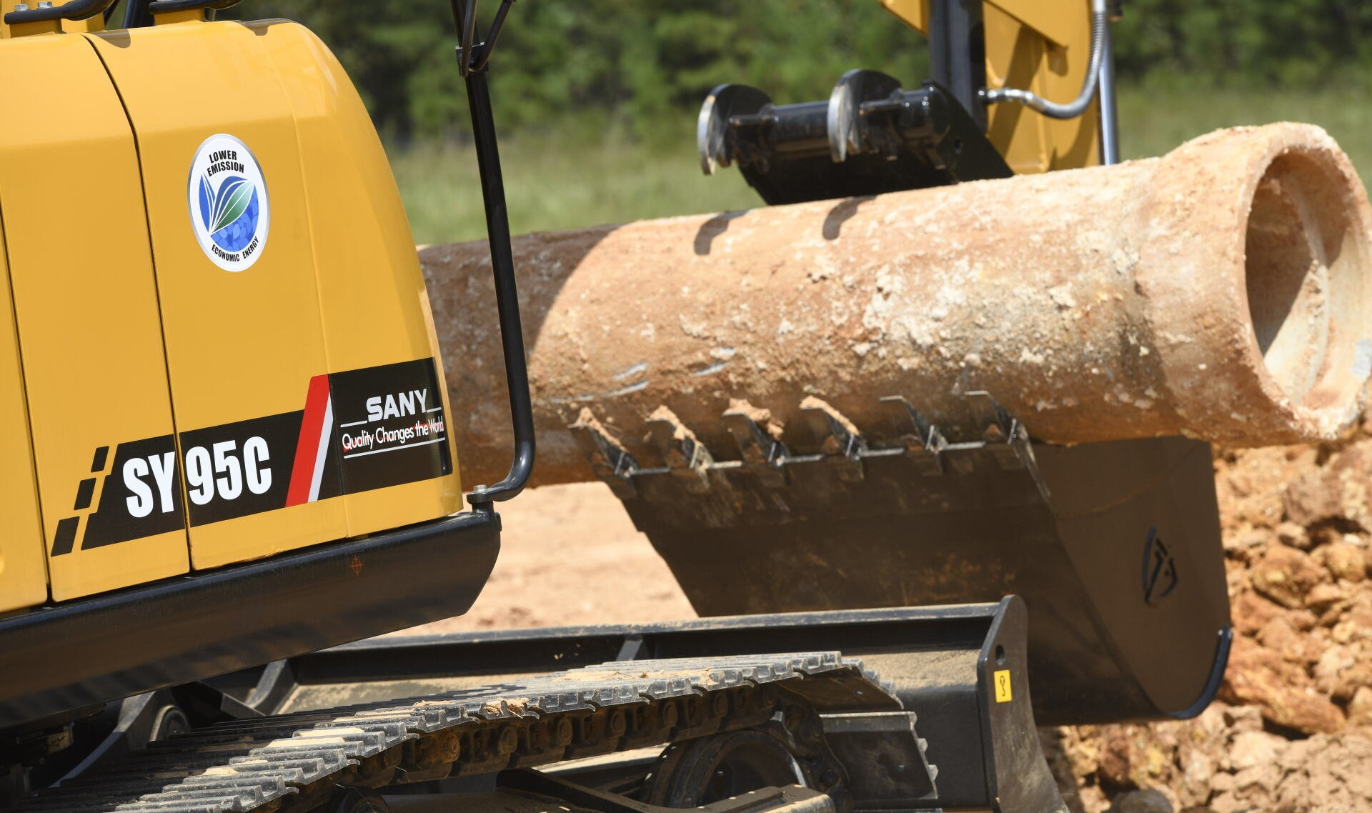 A yellow excavator is carrying a large pipe in its bucket.
