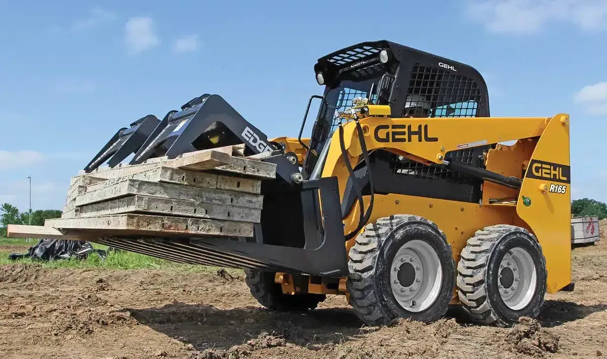 A yellow and black new holland skid steer loader is driving down a dirt road.