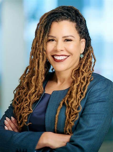 A woman with dreadlocks and a blue jacket is smiling with her arms crossed.