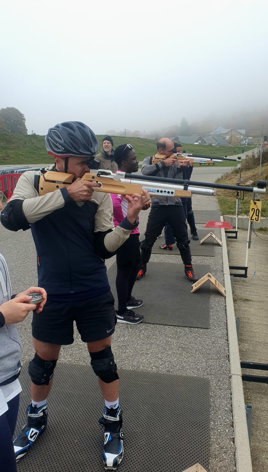 Personne en bottes de ski pointant un fusil sur un champ de tir, avec d'autres en arrière-plan, décor extérieur brumeux.