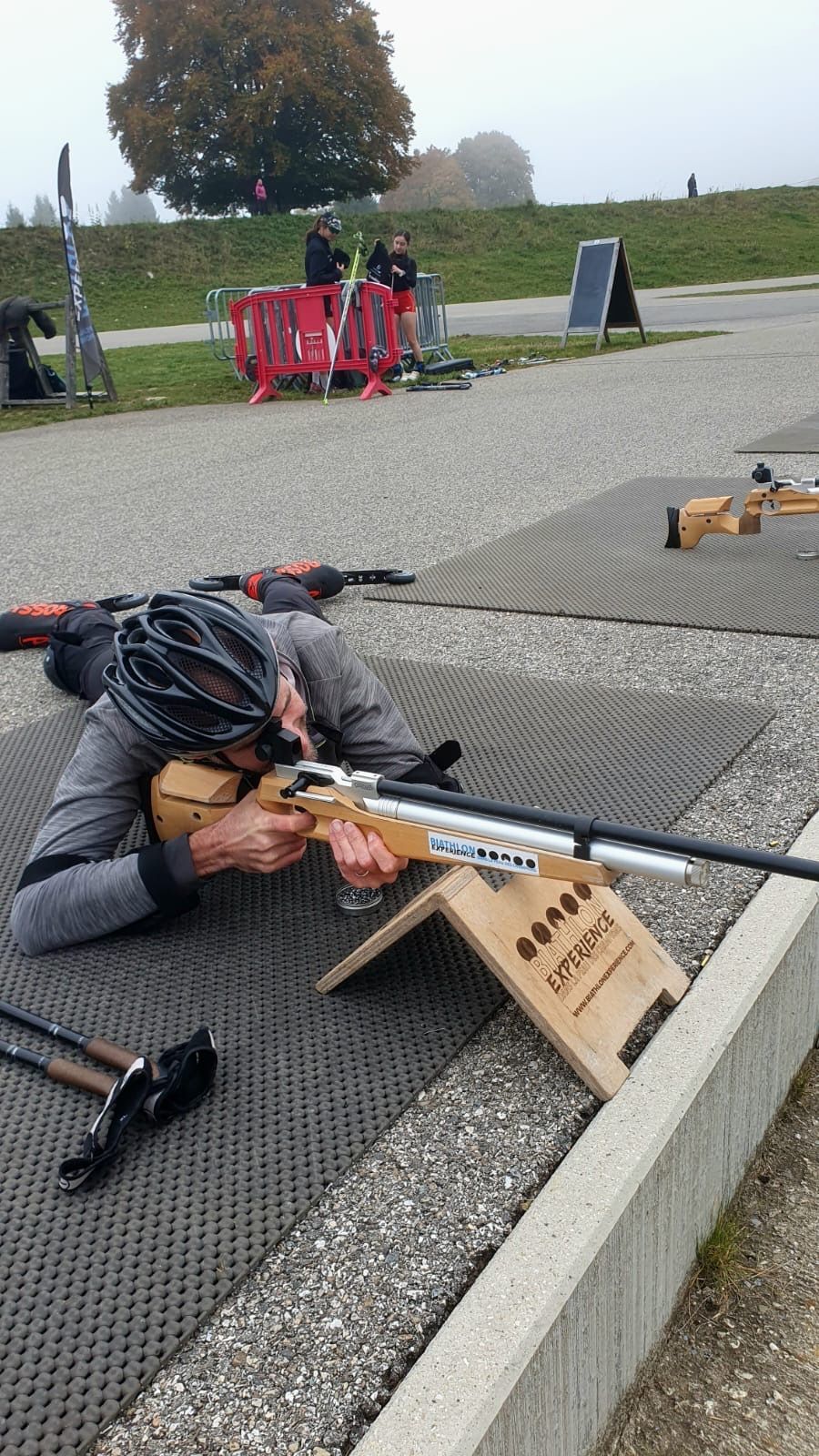 Un homme vêtu d'une veste grise pointe un fusil vers un stand de tir, allongé sur le ventre. Extérieur, ciel couvert.
