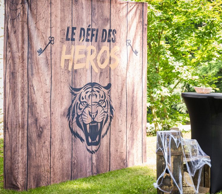 Fond en bois avec inscription « Le Défi des Héros », tête de tigre et clés. Herbe verte, arbres et table visibles.