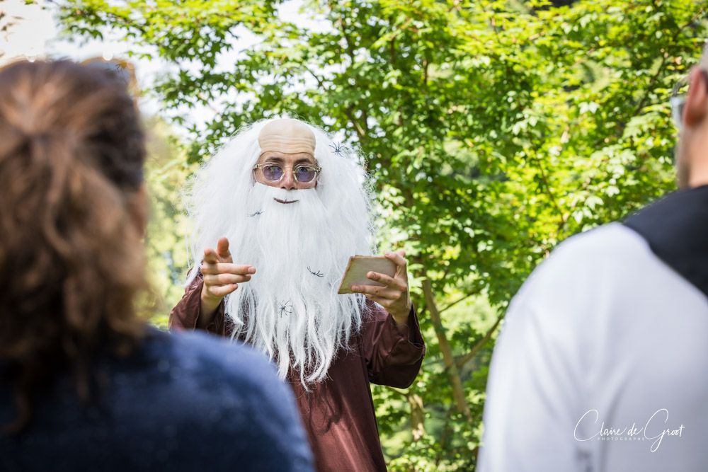 Une personne en costume de sorcier fait un geste, face à deux personnes à l'extérieur avec des arbres en arrière-plan.