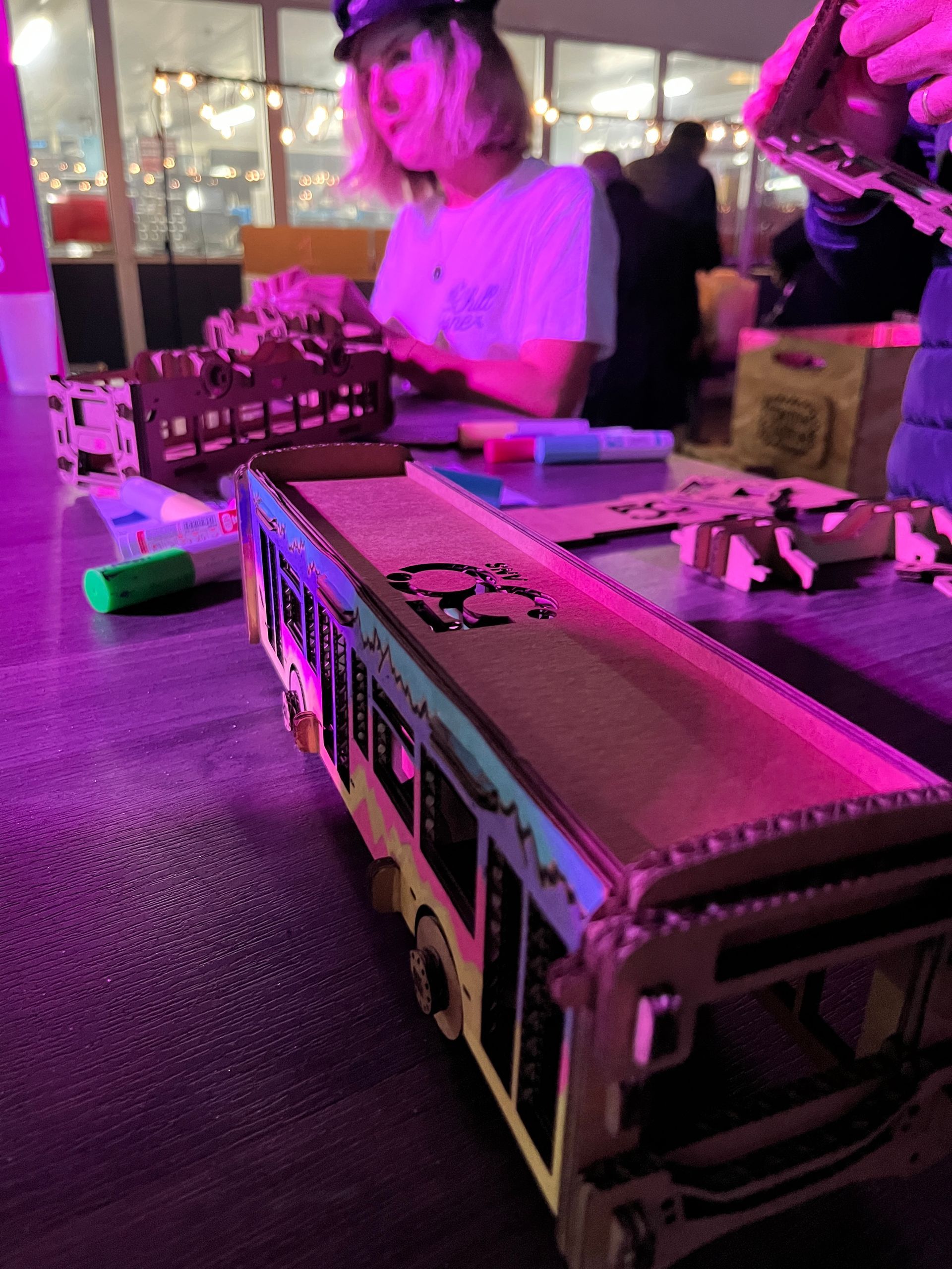 Une table avec un modèle de bus en arrière-plan ; une personne à l'arrière-plan assemble des structures en carton.