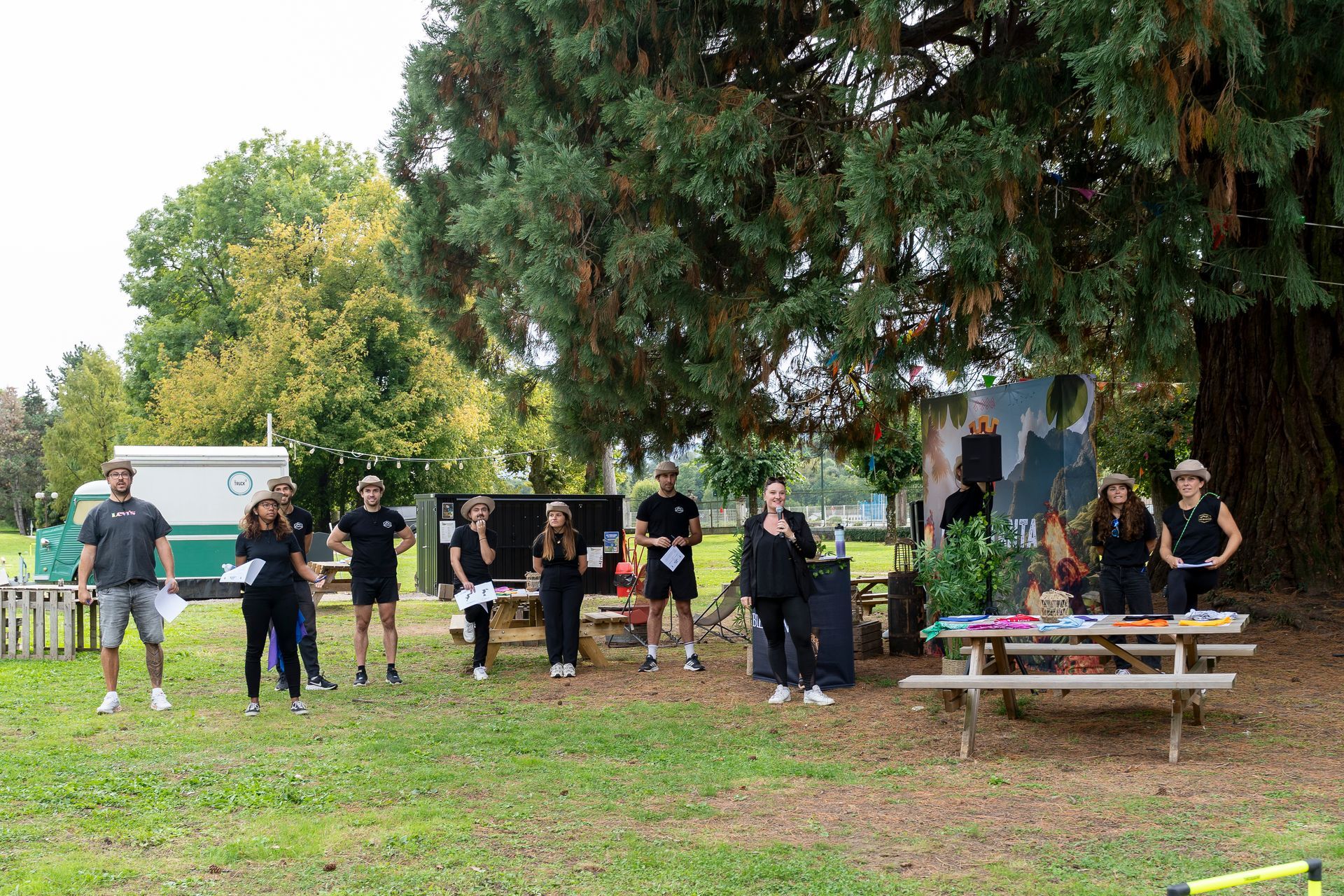 Un groupe de personnes se tenant à l'extérieur, probablement lors d'un événement, près de tables de pique-nique et d'un grand arbre.