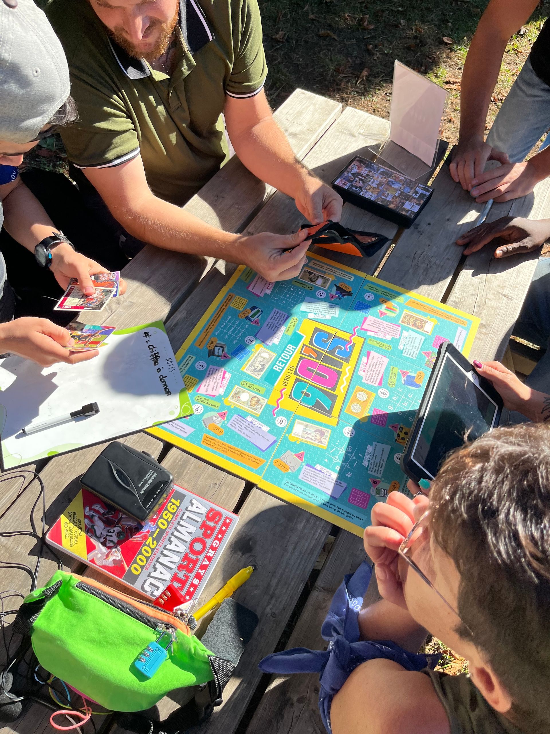 Des gens jouent à un jeu de société en plein air. Les mains se tendent vers les pièces du jeu sur un plateau coloré.