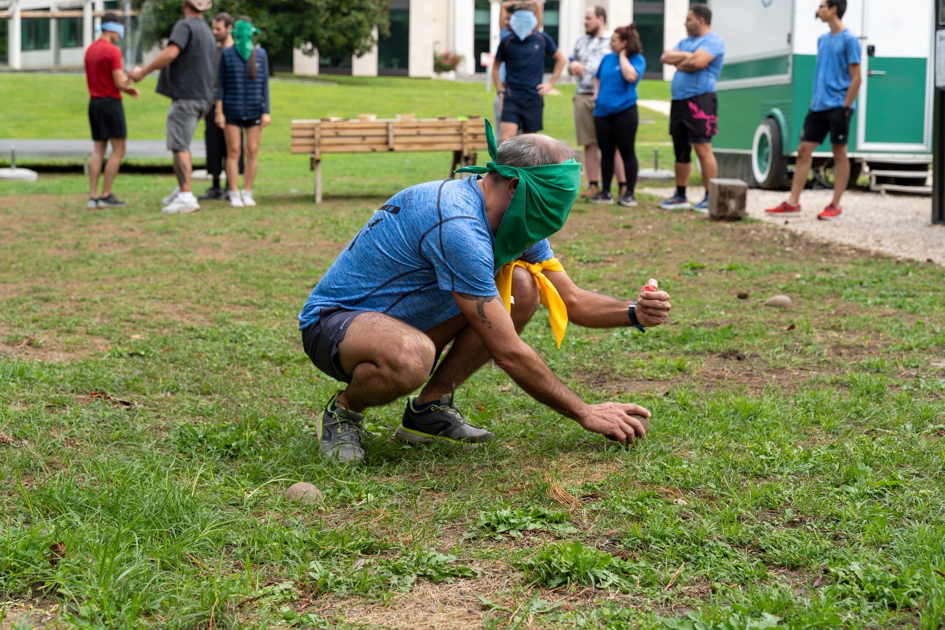 Un homme, les yeux bandés, accroupi dans l'herbe, tente d'attraper quelque chose. D'autres personnes l'observent dans un parc.