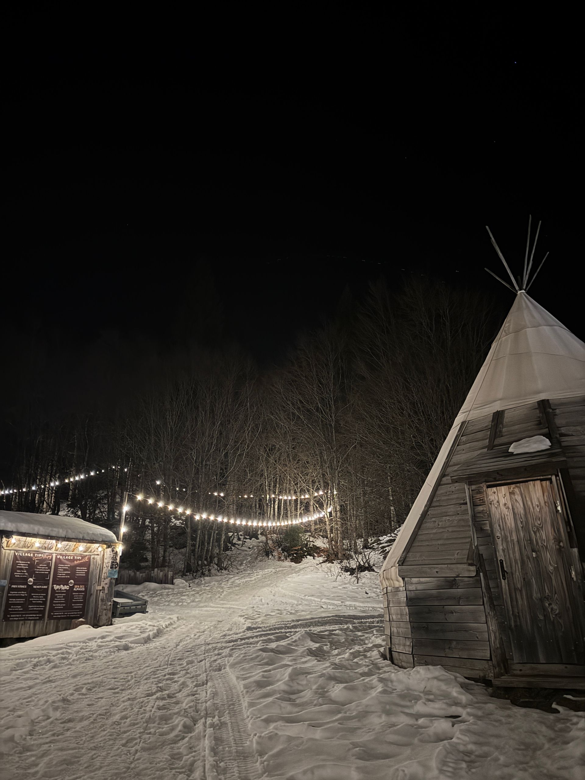 Scène de nuit enneigée avec tipi, structure en bois et guirlandes lumineuses ; arbres en arrière-plan.