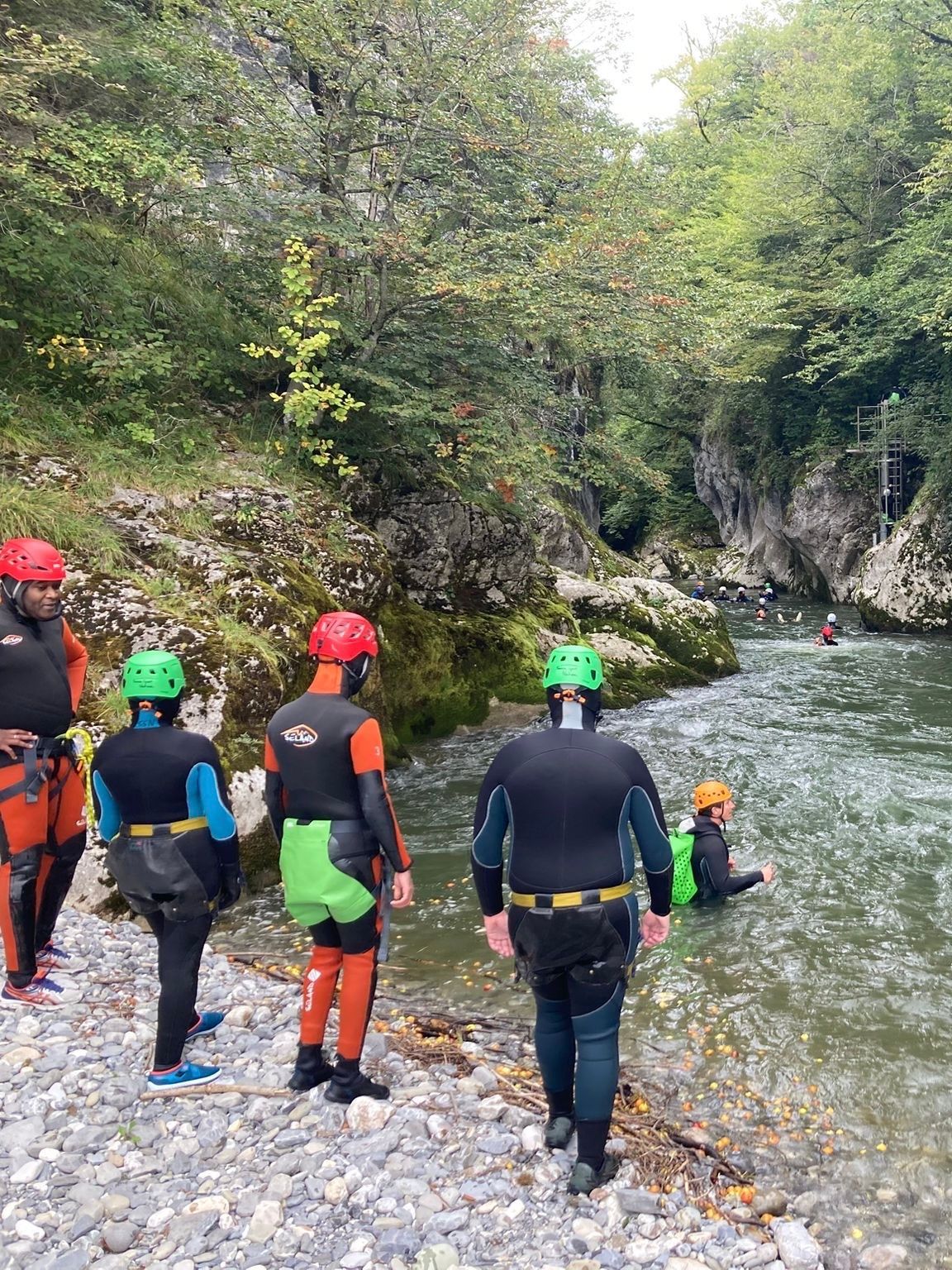 Des personnes en combinaison de plongée se préparent à entrer dans une rivière, entourée d'une rive rocheuse et d'arbres.