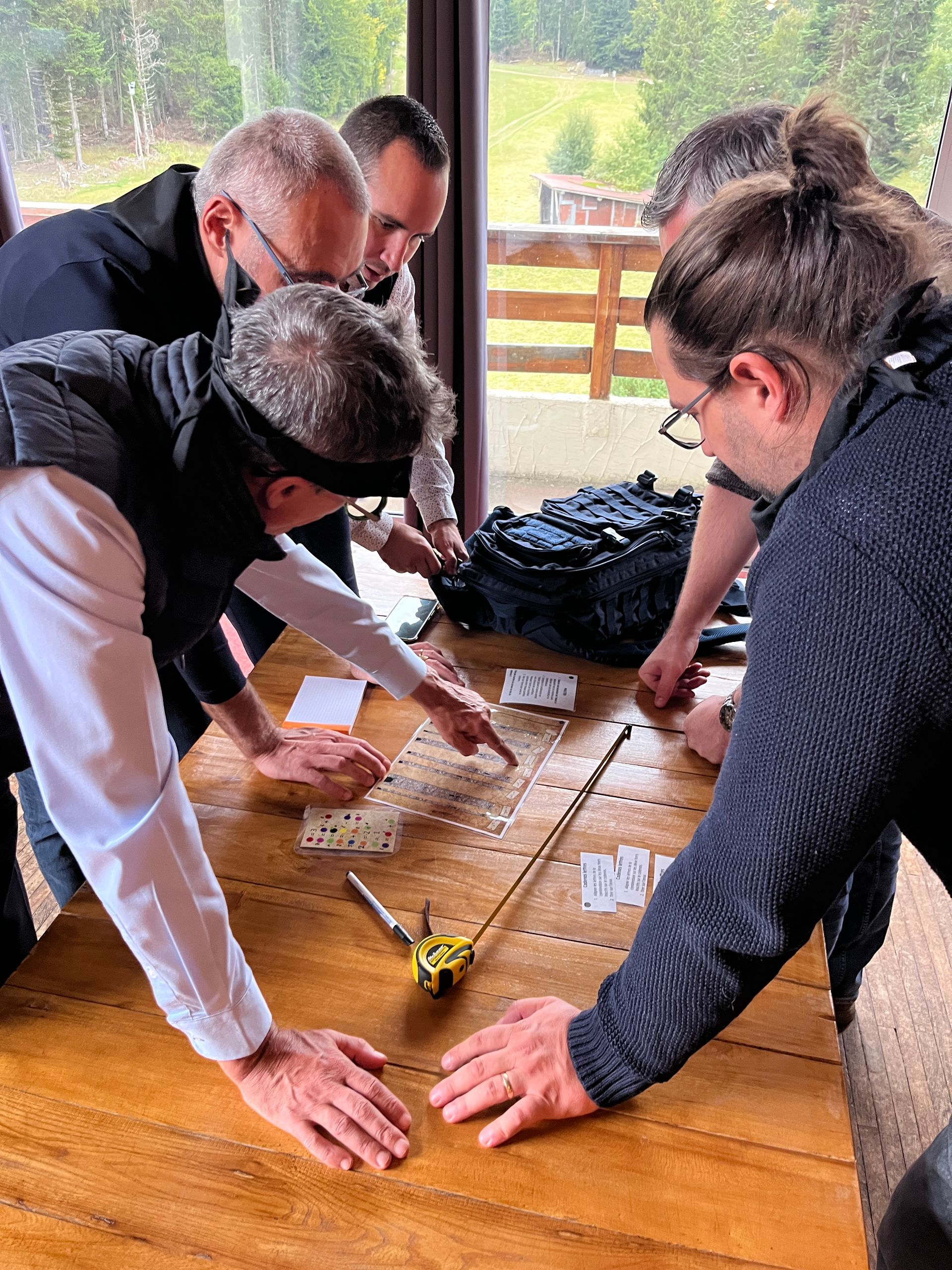 Cinq personnes à une table, dont une aux yeux bandés, pointant des cartes. Ambiance nature, travail d'équipe.