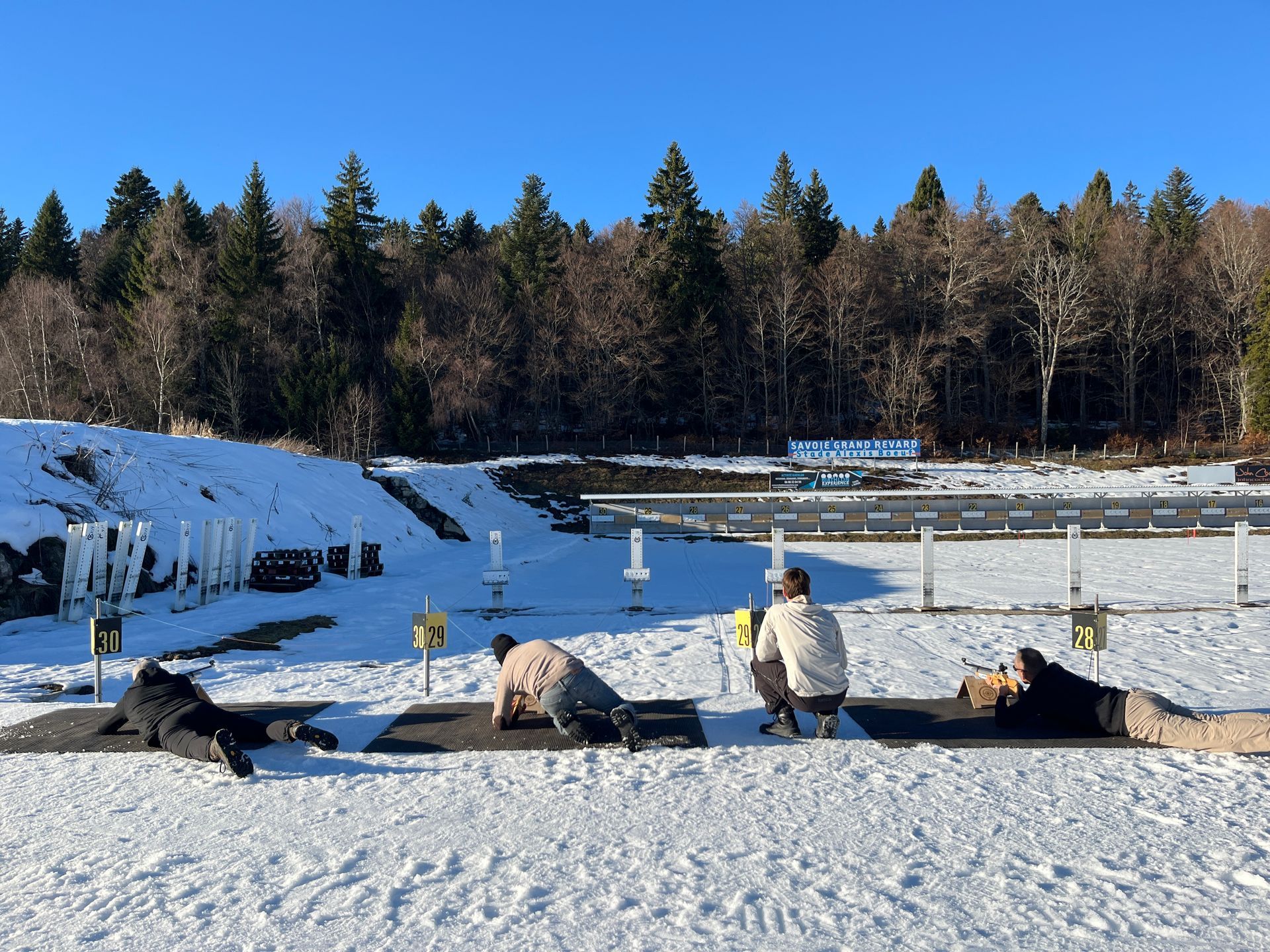 Des personnes tirent au fusil sur un stand de tir extérieur enneigé, avec des cibles au loin et une forêt en arrière-plan.