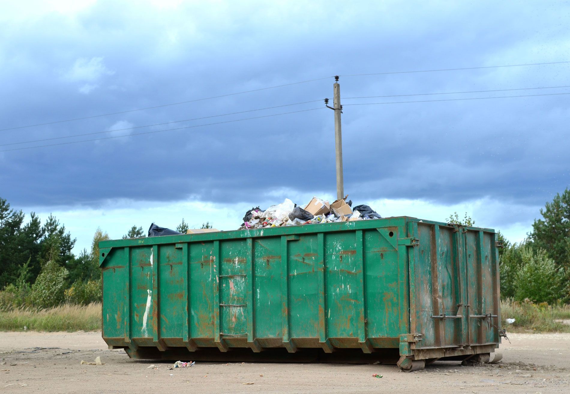 A large green dumpster filled with trash is sitting in the middle of a dirt field.