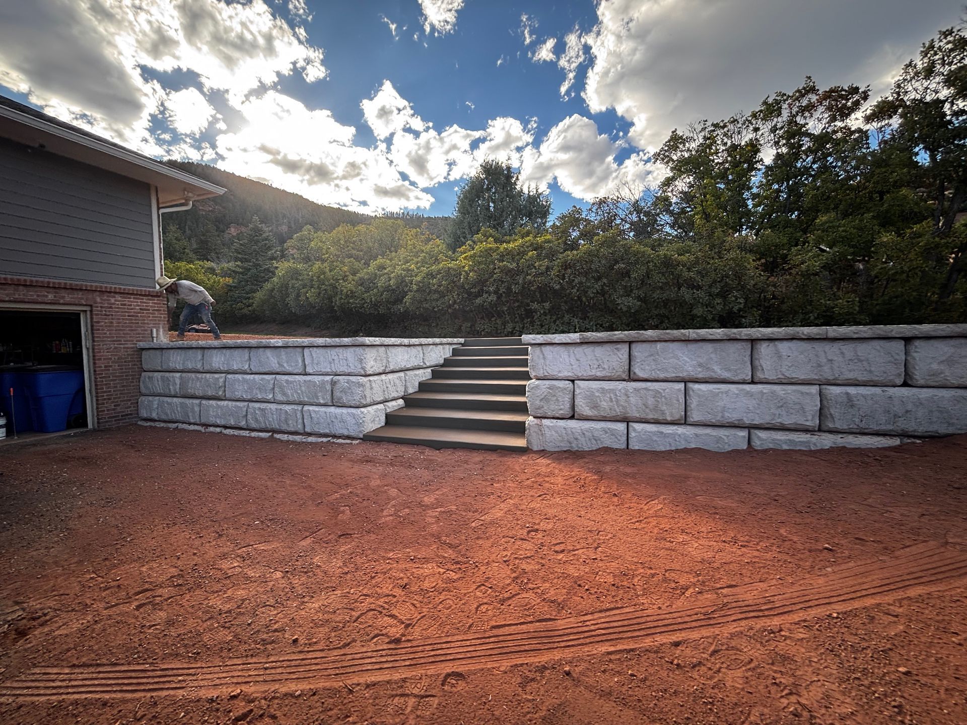 Stone steps in a red dirt courtyard beside a house, with a low retaining wall and cloudy sky.