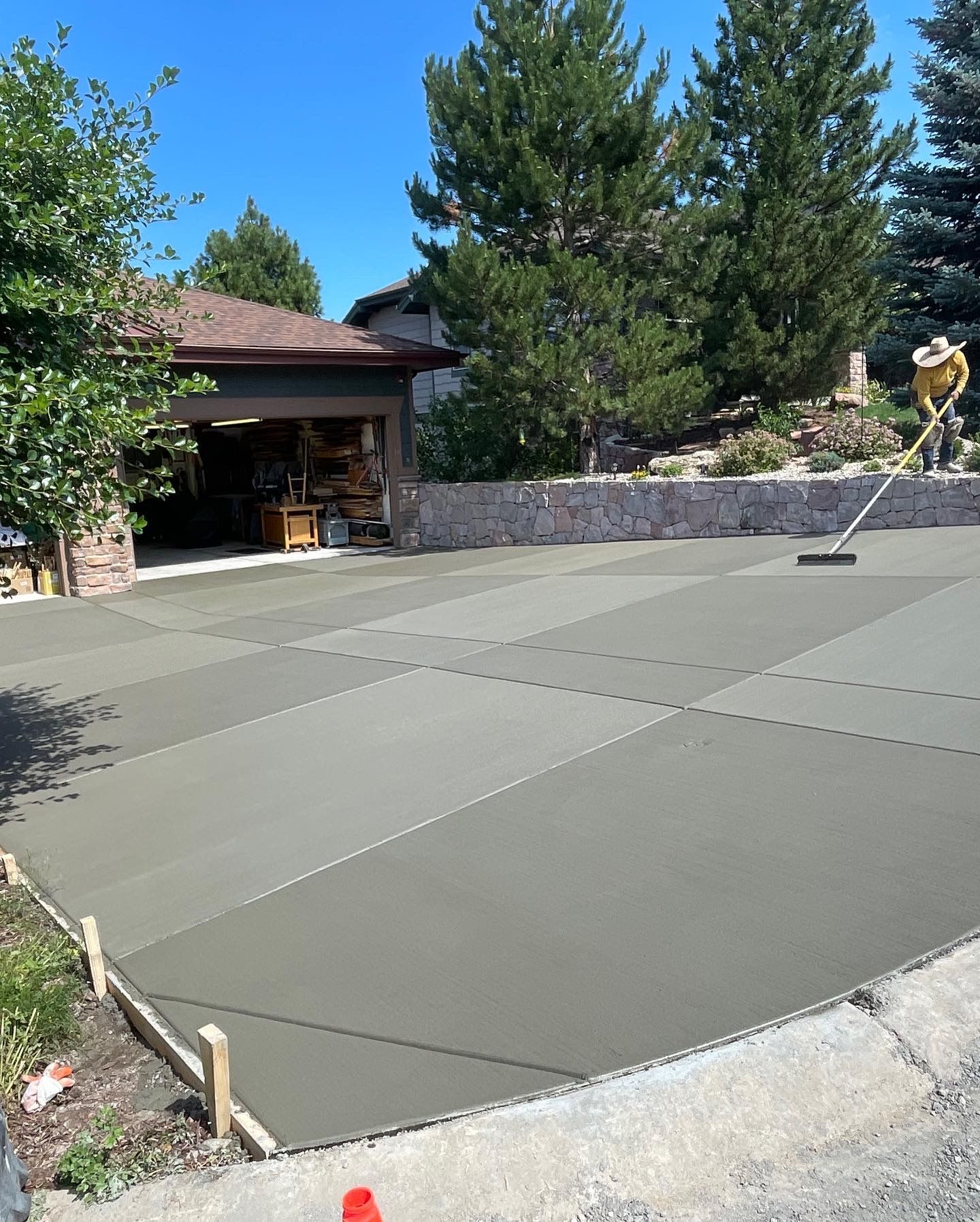 Freshly poured concrete driveway beside a house and stone wall, with trees in the background