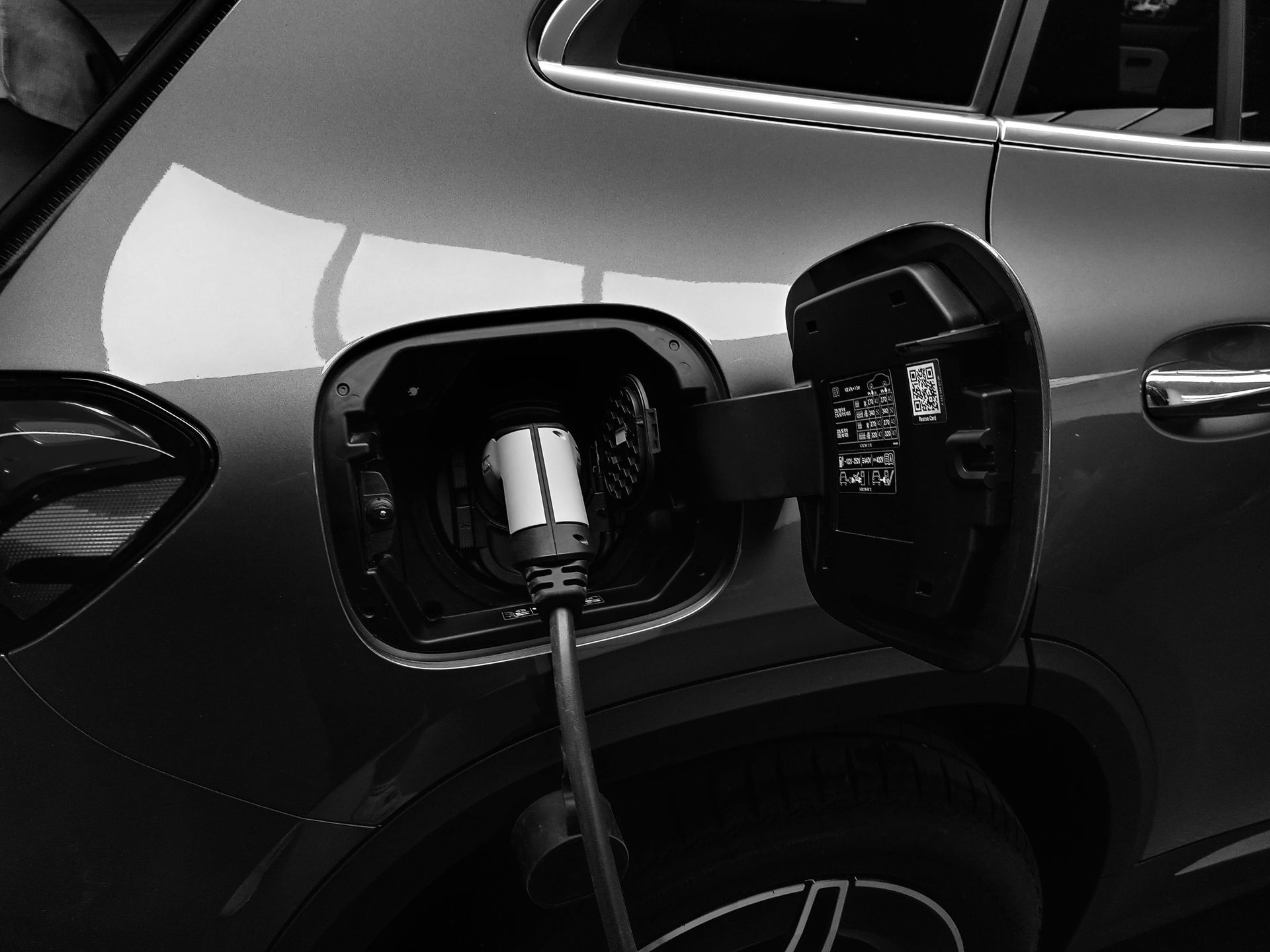 Electric vehicle charging, black and white close-up. Plug inserted into car's charging port with open door.