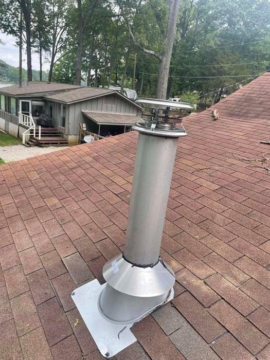 Metal chimney on a brown shingled roof with a house and trees in the background.