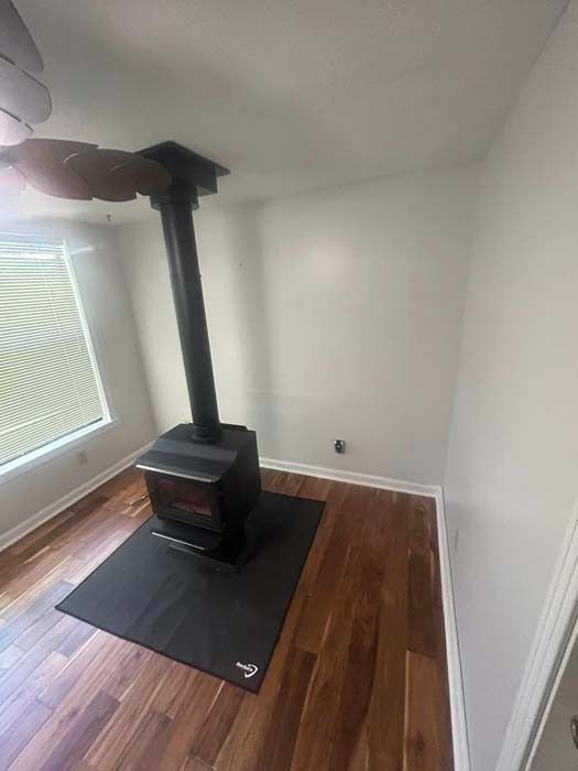 Wood-burning stove with black pipe in a room with wood flooring and a window with blinds.