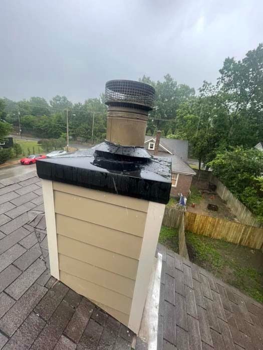 Chimney on a roof with a rain cap, tan siding, gray shingles, and trees in the background.