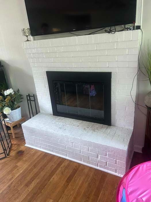 White brick fireplace with black insert and TV above. Wood floors and potted plants surround.