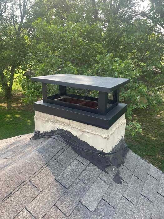 Chimney with black cap on a roof with gray shingles, trees in the background.