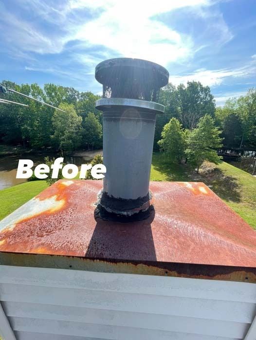 A rusty rooftop with a gray chimney and cap against a bright sky with trees and a river in the background.