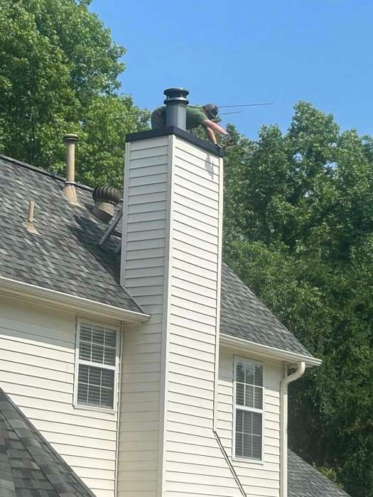 Person on a roof repairs a chimney. Tall white chimney with black cap, person in green shirt.