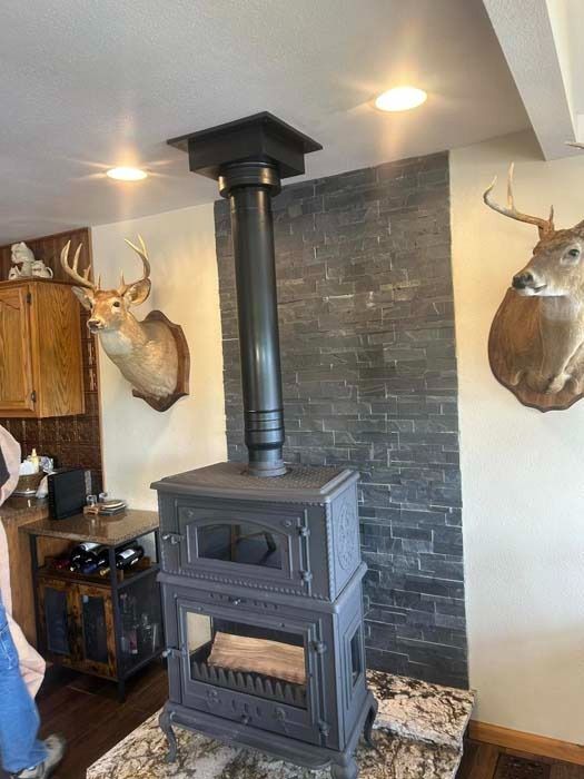 A black wood-burning stove with a vertical pipe against a stone-tiled wall, flanked by deer head mounts.