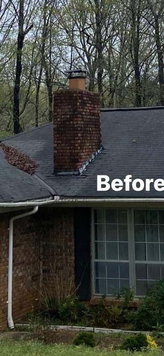 Brick chimney on a house with a dark roof and white trim. Green foliage surrounds the house.