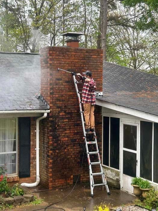 Person power washing a brick chimney from a ladder on a residential roof.