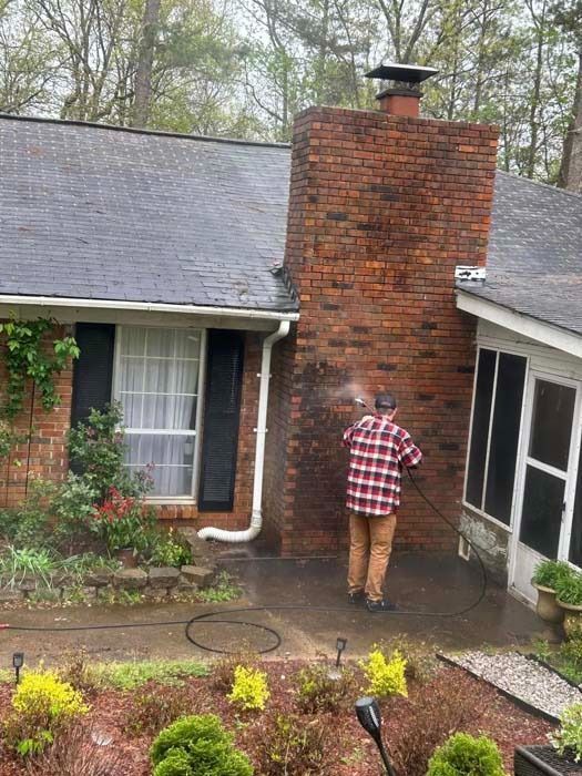 Person pressure washing a red brick chimney on a house; wearing plaid shirt, standing outside.