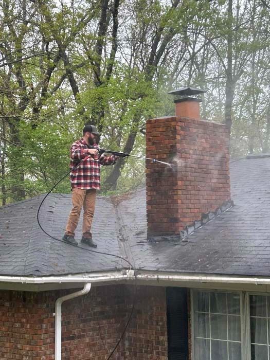 Man on a roof power washing a brick chimney; trees in background.