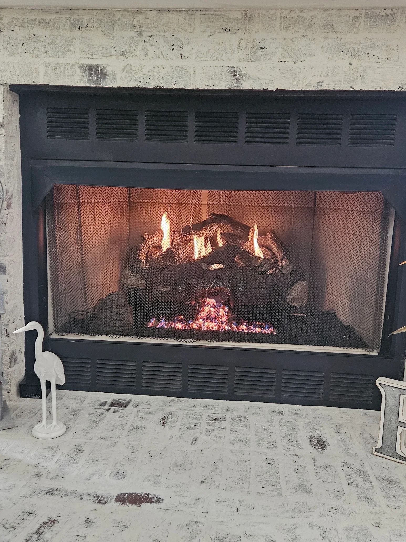 Fireplace with burning logs behind a black mesh screen, inside a light gray brick frame.