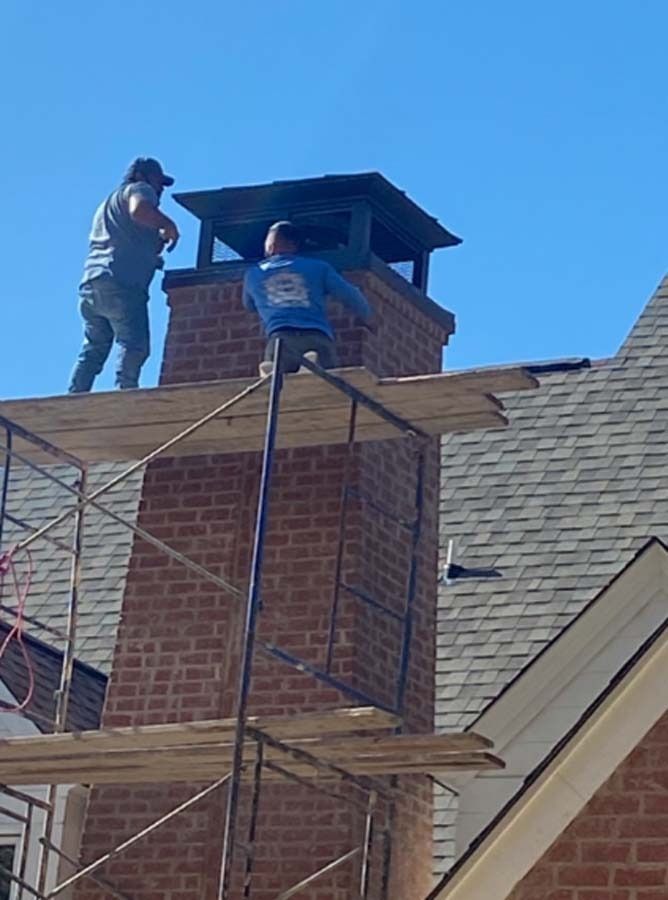 Two workers on scaffolding repairing a brick chimney. Bright blue sky, gray shingles.