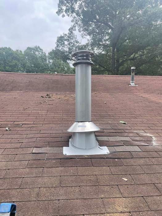 Gray metal chimney on a brown shingled roof, with trees in the background on a cloudy day.