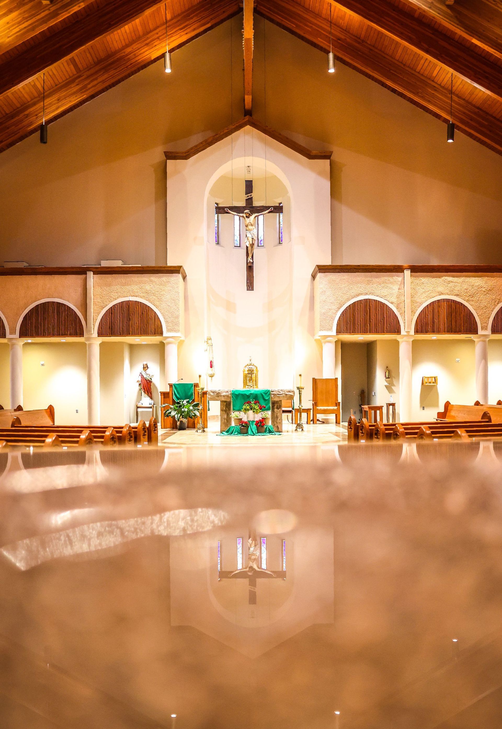 Interior view of a St. Katharine Drexel Catholic Church with wooden vaulted ceiling. The altar, adorned with a green cloth, is centered with a crucifix hanging above.