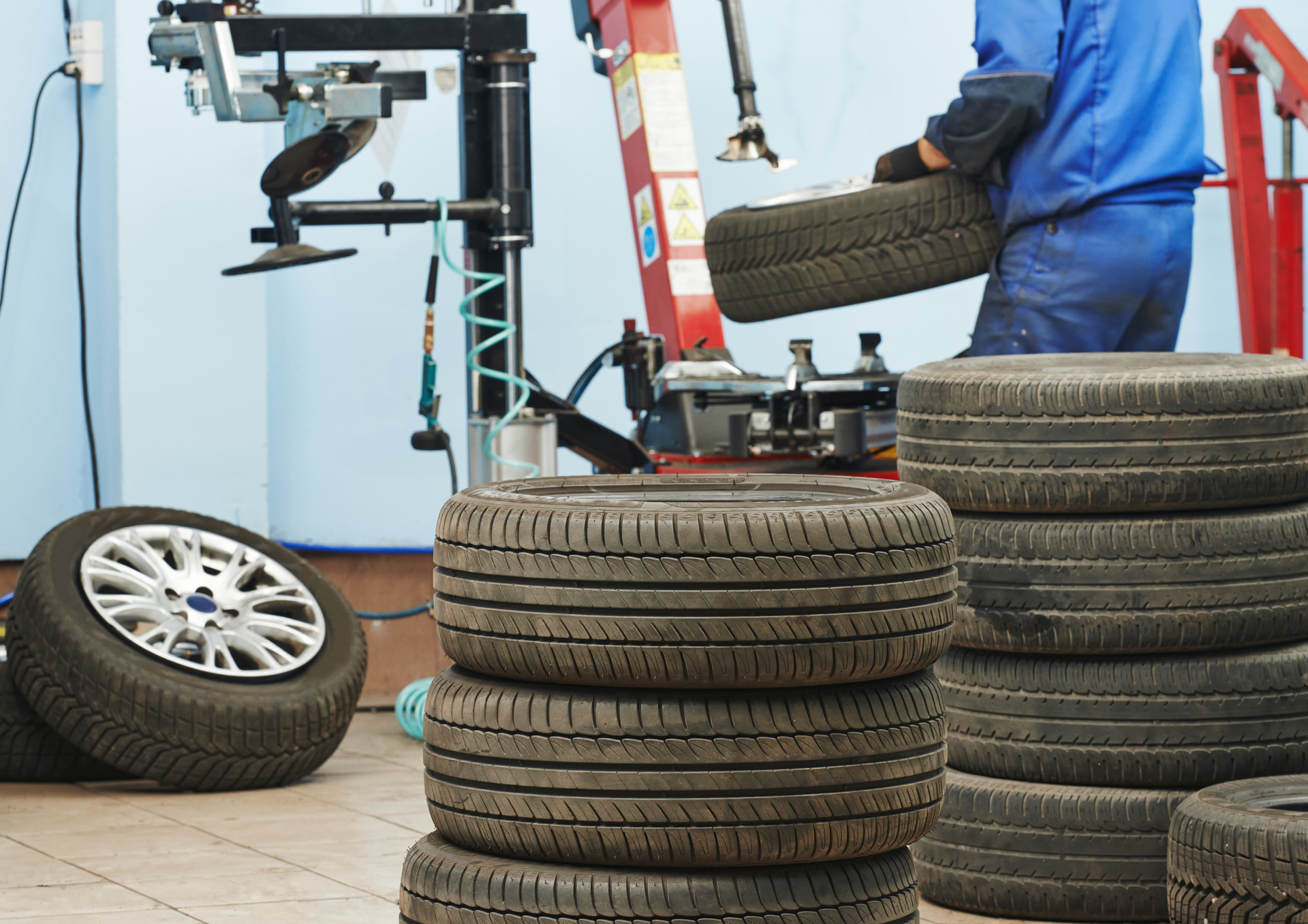 A mechanic in a blue uniform working at a tire changing machine in a repair shop with several stacks of tires.