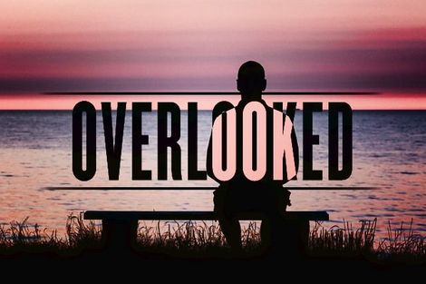 This is a photograph of a person sitting on a bench alone and the over text says Overlooked.
