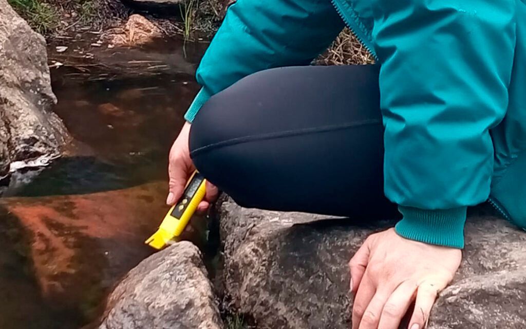 Una persona está arrodillada sobre una roca junto a un arroyo sosteniendo un objeto amarillo.