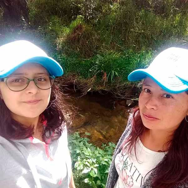 Dos mujeres con gorras de béisbol y gafas están paradas una al lado de la otra frente a un arroyo.